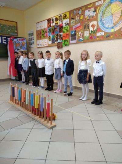 Dzieci stoją obok siebie w jednym szeregu. Przed uczniami dekoracja w kształcie ołówków. Za uczniami na ścianie wiszą piękne, jesienne dekoracje.