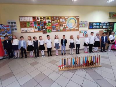 Dzieci stoją w jednym szeregu, przed nimi znajduje się dekoracja w postaci płotka, na którym znajdują się ołówki. Za dziećmi jesienne dekoracje na tablicach.