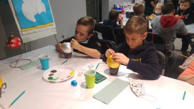 Dwaj chłopcy siedzą przy stole w Centrum Dziedzictwa Szkła i malują farbami bombki choinkowe.