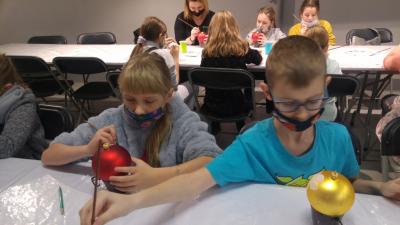 Dzieci siedzą przy stolikach i ozdabiają farbami bombki choinkowe.