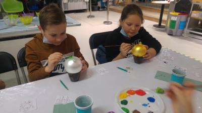 Dzieci siedzą przy stole w Centrum Dziedzictwa Szkła i malują farbami bombki choinkowe.