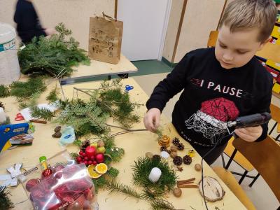 Uczniowie klasy IVb uczestniczą w warsztatach z tworzenia stroików świątecznych. Na stołach leżą gałązki jodły, bombki. Uczniowie stoją wokół stołu i wykonują stroiki