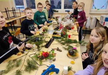 Uczniowie klasy IVb uczestniczą w warsztatach z tworzenia stroików świątecznych. Na stołach leżą gałązki jodły, bombki. Uczniowie stoją wokół stołu i wykonują stroiki