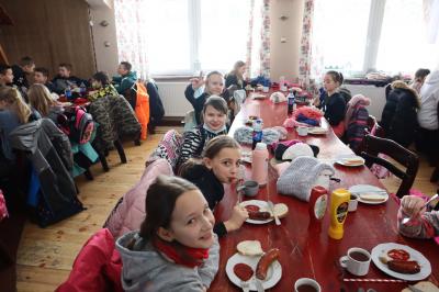 Dzieci siedzą w karczmie przy stołach i spożywają kiełbaski oraz piją herbatę.