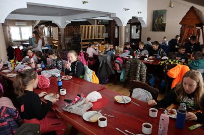Dzieci siedzą w karczmie przy stołach i spożywają kiełbaski oraz piją herbatę.