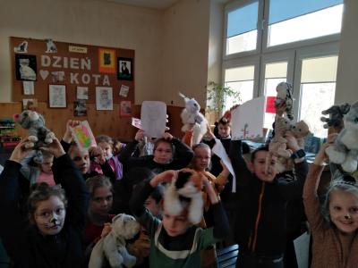 Zdjęcie przedstawia uczniów, którzy trzymają w rękach nad głowami, przyniesione ze sobą pluszowe koty lub wykonane przez siebie rysunki kotów. Niektórzy z nich mają nawet pomalowane twarze lub dorysowane kocie wąsy.