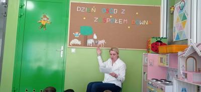 Nauczyciel siedzi na krzesełku przed tablicą korkową , czyta opowiadanie dzieciom i wskazuje sylwety zwierząt występujących w tym utworze.
