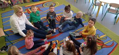 Dzieci i nauczyciel siedzą w kole na dywanie w sali ze zdjętym obuwiem zastępczym. Każdy na stopach ma inną skarpetkę.