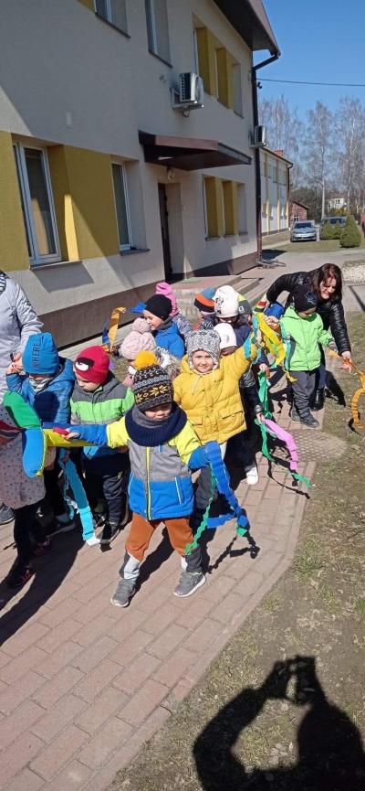 Dzieci idą na spacer koło budynku szkoły i trzymają w rękach kolorowe bibułkowe wstążki.