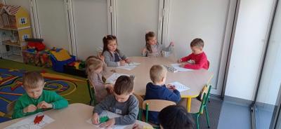 Dzieci siedzą przy stolikach w sali i kolorują kredkami pastelowymi szablony skarpetek.