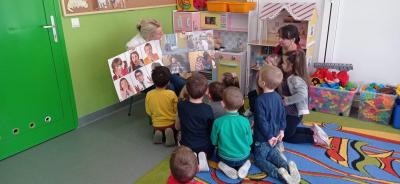 Nauczyciel siedzi na krzesełku przed dziećmi skupionymi wokół niego i pokazuje ilustrację mimiki twarzy i zachowań ludzi  w różnym wieku i w różnych stanach emocjonalnych.