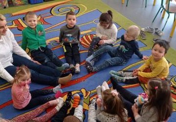 Dzieci i nauczyciel siedzą w kole na dywanie w sali ze zdjętym obuwiem zastępczym. Każdy na stopach ma inną skarpetkę.