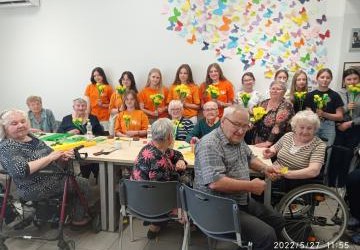 Przy wspólnym stole siedzą seniorzy z Lokalnego Ośrodka Wsparcia i Aktywizacji w Libuszy. Przy ścianie stoją wolontariusze SKW ubrani w pomarańczowe podkoszulki trzymając w rękach wykonane własnoręcznie żonkile.