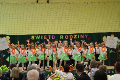 Przedszkolaki z grupy Leśne Duszki występują na scenie - w tle napis święto rodziny. Przed sceną pięknie udekorowane kwiaty w doniczkach.