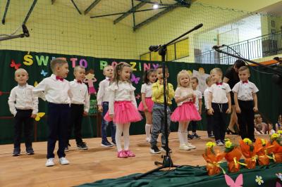 Przedszkolaki z grupy Sówki występują na scenie - w tle napis święto rodziny. Przed sceną pięknie udekorowane kwiaty w doniczkach.