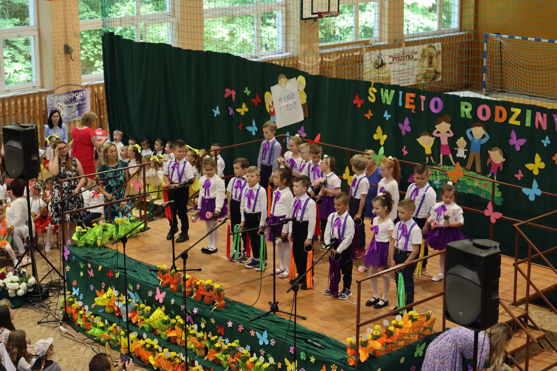 Przedszkolaki z grupy Skrzatów występują na scenie - w tle napis święto rodziny. Przed sceną pięknie udekorowane kwiaty w doniczkach.