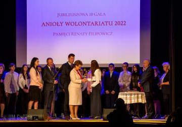 Opiekun SKW odbiera z rąk strarosty powiatu gorlickiego p.Marii Gubała statuetkę Anioły Wolontariatu 2022. Na scenie stoją również dyrektor ZSP w Libuszy p.Aleksander Kalisz wraz z wolontariuszami SKW.