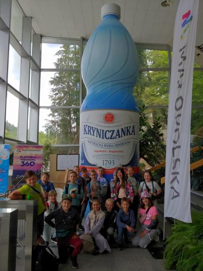 Grupa dzieci pozuje do zdjęcia na tle dużej butelki z napisem Kryniczanka