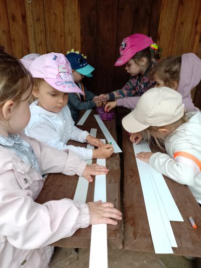 Dzieci siedzą przy drewnianych stolikach i kolorują paski papieru, z których powstaną opaski indiańskie.  Na stoliku w pudełku leżą kredki woskDzieci z zaangażowaniem wykonują pracę.