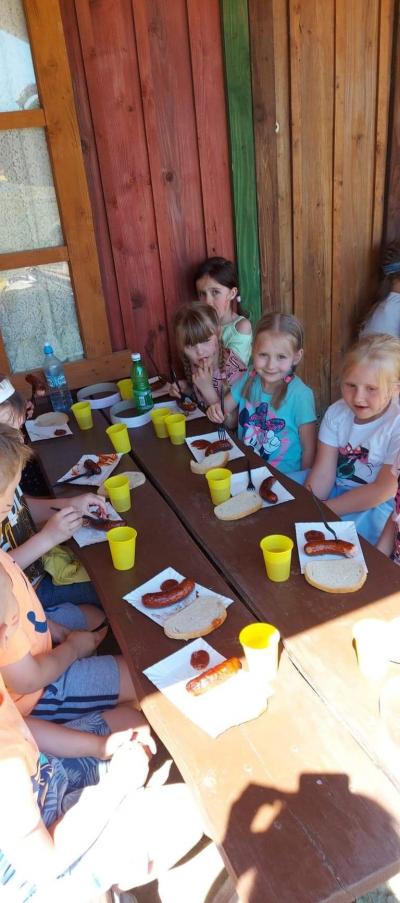 Na zdjęciu widać dzieci siedzące na ławkach przy drewnianych stolikach. Na stole znajdują się papierowe talerzyki z kiełbaską i chlebem oraz keczupem, a także kubeczki jednorazowe z herbatą. Dzieci spożywają posiłek. W tle widać ścianę drewnianego zabu