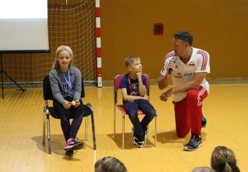 Marcin Prus ubrany w strój sportowy w narodowych barwach, trzyma mikrofon w dłoni zadając pytania dwóm uczniom Zespołu Szkolno - Przedszkolnego w Libuszy. Spotkanie w hali sportowej. Uczniowie siedzą na krzesełkach.