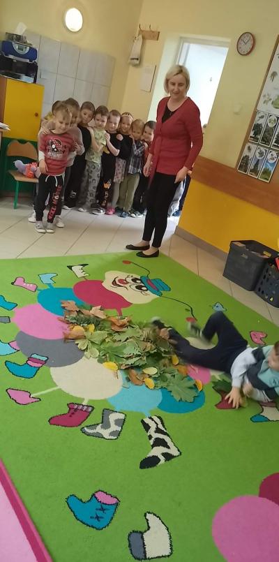 Dzieci stoją w szeregu przed dywanem na którym zgromadzony jest stos liści. Chłopiec skacze w liście i przewraca się. Nauczyciel stoi obok nadzorując zabawę.