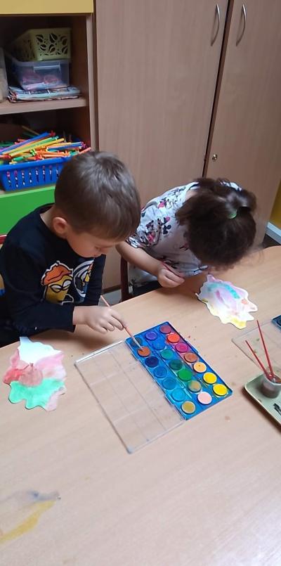 Przy stoliku przedszkolnym siedzą dziewczynka i chłopiec, którzy malują farbami plakatowymi sylwety liścia dębu.