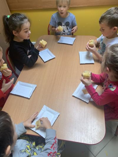 Sześcioro dzieci siedzi przy stoliku i jedzą biszkopt z jabłkami z papierowych tacek.