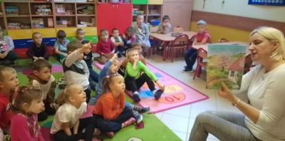 Dzieci siedzą w sali przedszkolnej na dywanie. Nauczyciel siedzi przed nimi i pokazuje obrazki ilustrujące powstawanie owocu jabłka od momentu zapylenia do dojrzałego owocu.