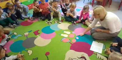 Dzieci siedzą w kole razem z nauczycielem na dywanie w sali przedszkolnej. Na dywanie rozłożone są sylwety trzech jabłek, dwóch gruszek i trzech śliwek. Nauczyciel zabiera jedną śliwkę, a dzieci przeliczają ilość sylwet owoców pozostałych na dywanie.