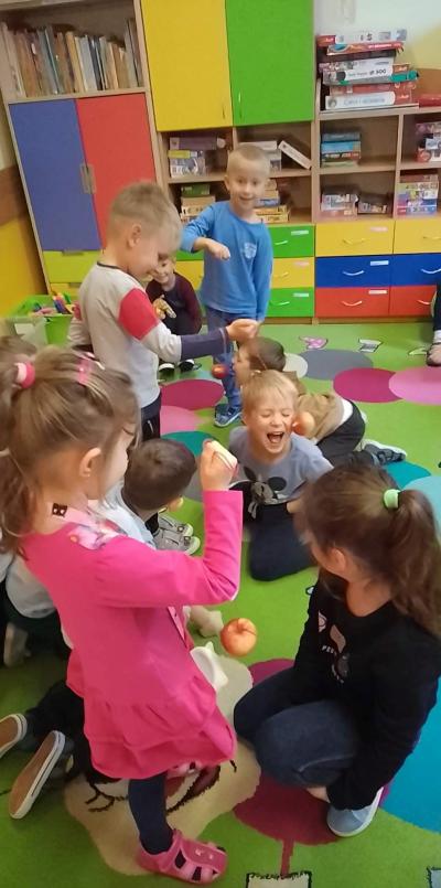 Troje dzieci stoi i trzyma w dłoniach jabłka zawiązane za ogonek na sznurku. Troje innych dzieci klęczy przed nimi i próbuje ugryźć jabłko bez trzymania go w dłoniach.