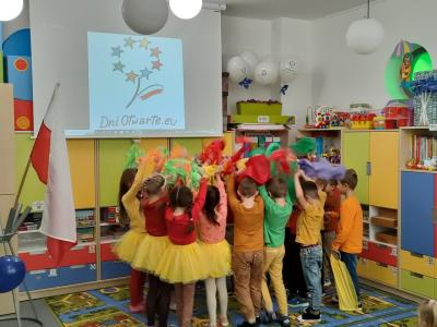 Dzieci w kółeczku twarzą do środka z rękami podniesionymi do góry, w rękach trzymają kolorowe chustki