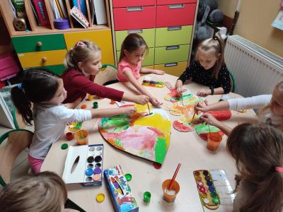 Dzieci siedzą przy stoliku, malują farbami duże, wycięte z kartonu liście.