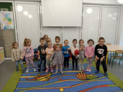 Grupa dzieci pozuje do zdjęcia, stoją na dywanie w sali przedszkolnej