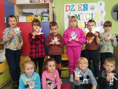 Wspólne zdjęcie dzieci z grupy Skrzaty wraz z pracami plastycznymi. W tle tablica z dekoracją z okazji Światowego Dnia Mycia Rąk.