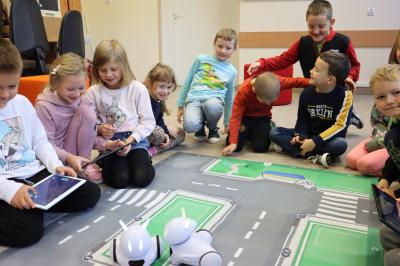Uczniowie klasy pierwszej uczestniczą w zajęciach z wykorzystaniem robotów Photonów. Dzieci siedzą dookoła maty i sterują robotami po uliczkach.