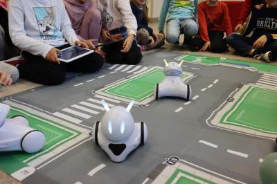 Uczniowie klasy pierwszej uczestniczą w zajęciach z wykorzystaniem robotów Photonów. Dzieci siedzą dookoła maty i sterują robotami po uliczkach.