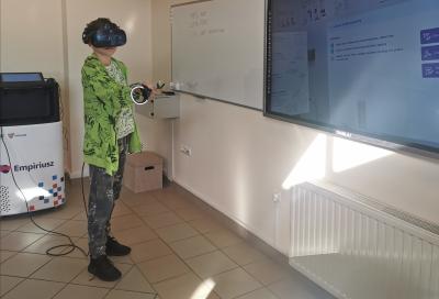 Uczeń obsługuje wirtualne laboratorium chemiczne Empiriusz. Za pomocą dżojstików przeprowadza doświadczenie chemiczne. Na głowie google VR. Reszta klasy obserwuje przeprowadzane doświadczenie.