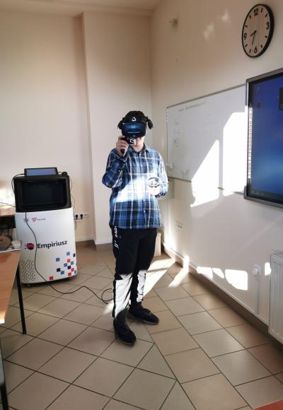 Uczeń obsługuje wirtualne laboratorium chemiczne Empiriusz. Za pomocą dżojstików przeprowadza doświadczenie chemiczne. Na głowie google VR. Reszta klasy obserwuje przeprowadzane doświadczenie.