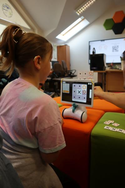 Dzieci z pomocą robota Photona rozgrywają pojedynek w grę kółko i krzyżyk podczas zajęć kończonych warsztaty związane ze sztuczną inteligencją. W pojedynku pomaga im nauczyciel.