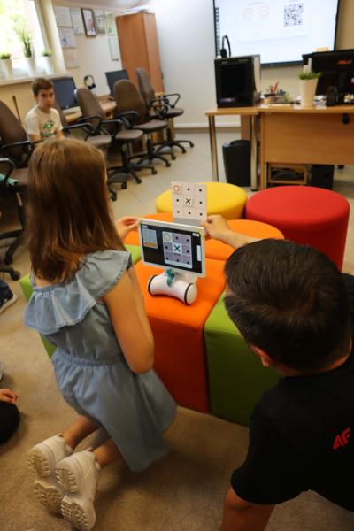 Dzieci z pomocą robota Photona rozgrywają pojedynek w grę kółko i krzyżyk podczas zajęć kończonych warsztaty związane ze sztuczną inteligencją. W pojedynku pomaga im nauczyciel.